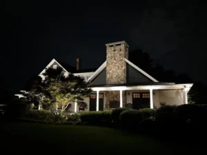 A nighttime scene of a house with a well-lit porch featuring white columns and seating area, surrounded by dark trees and shrubs, enhancing the architectural beauty.
