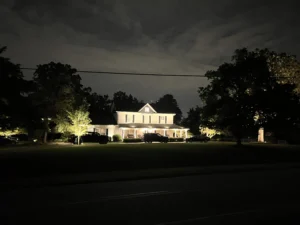 A nighttime view of a house with accent lighting on its porch and stone chimney, surrounded by illuminated bushes and trees, creating a cohesive outdoor scene.